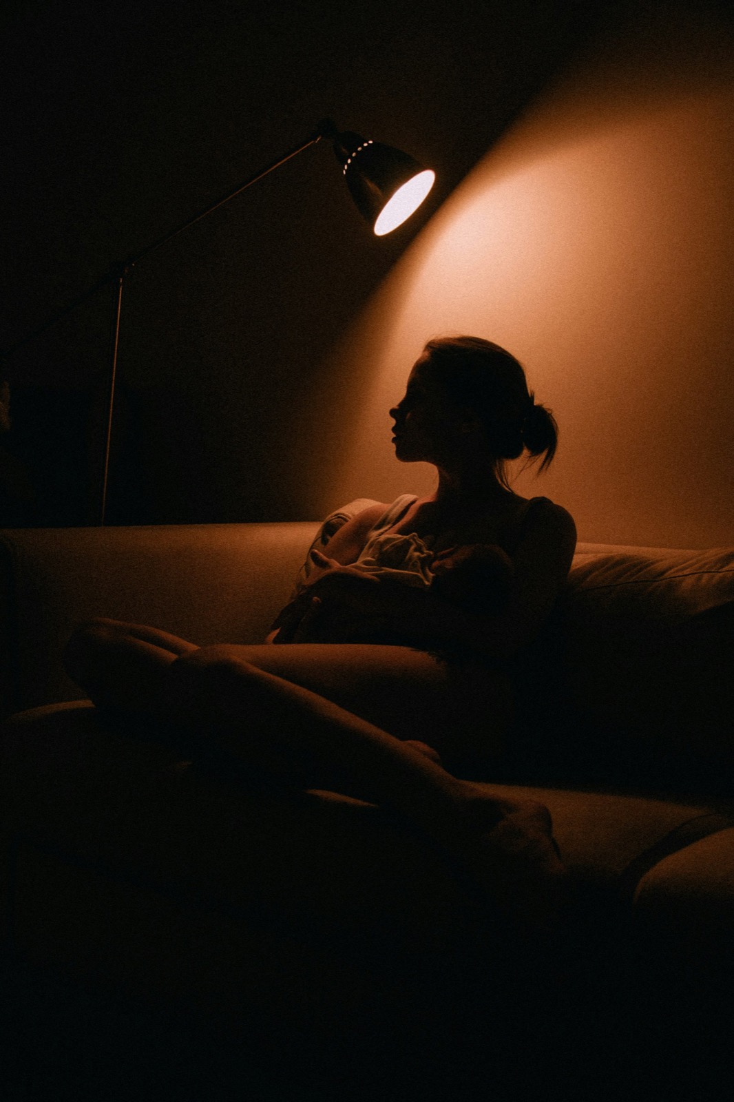 A woman resting on a sofa in a dim room lit by one lamp
